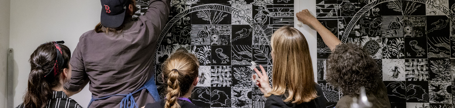 Five staff members hang up a large black and white print showing many small linocut squares.